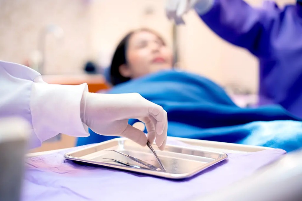 Gloved hand selecting a dental instrument from a tray with a patient in the background during in-house treatment